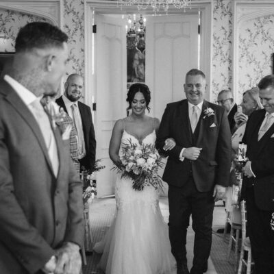 Black and white photo of bride walking down isle at Rowton Castle Shropshire with Father on arm, groom can be seen looking down towards the bride Shropshire Wedding Photographer